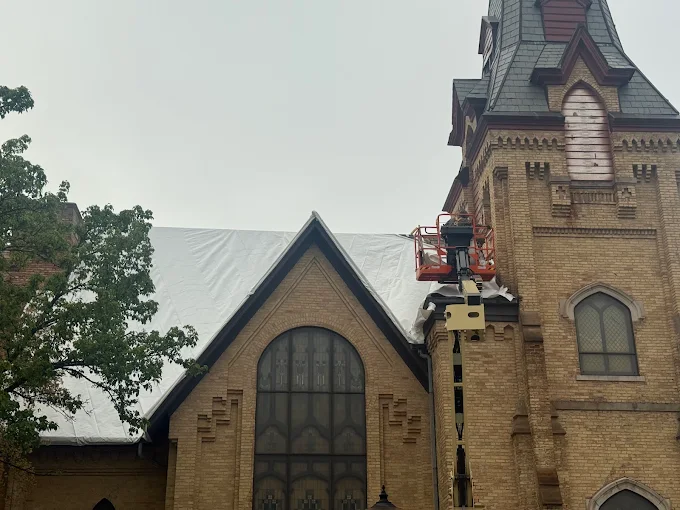Historic brick church with a white protective tarp over the main roof and a red boom lift raised against the steeple