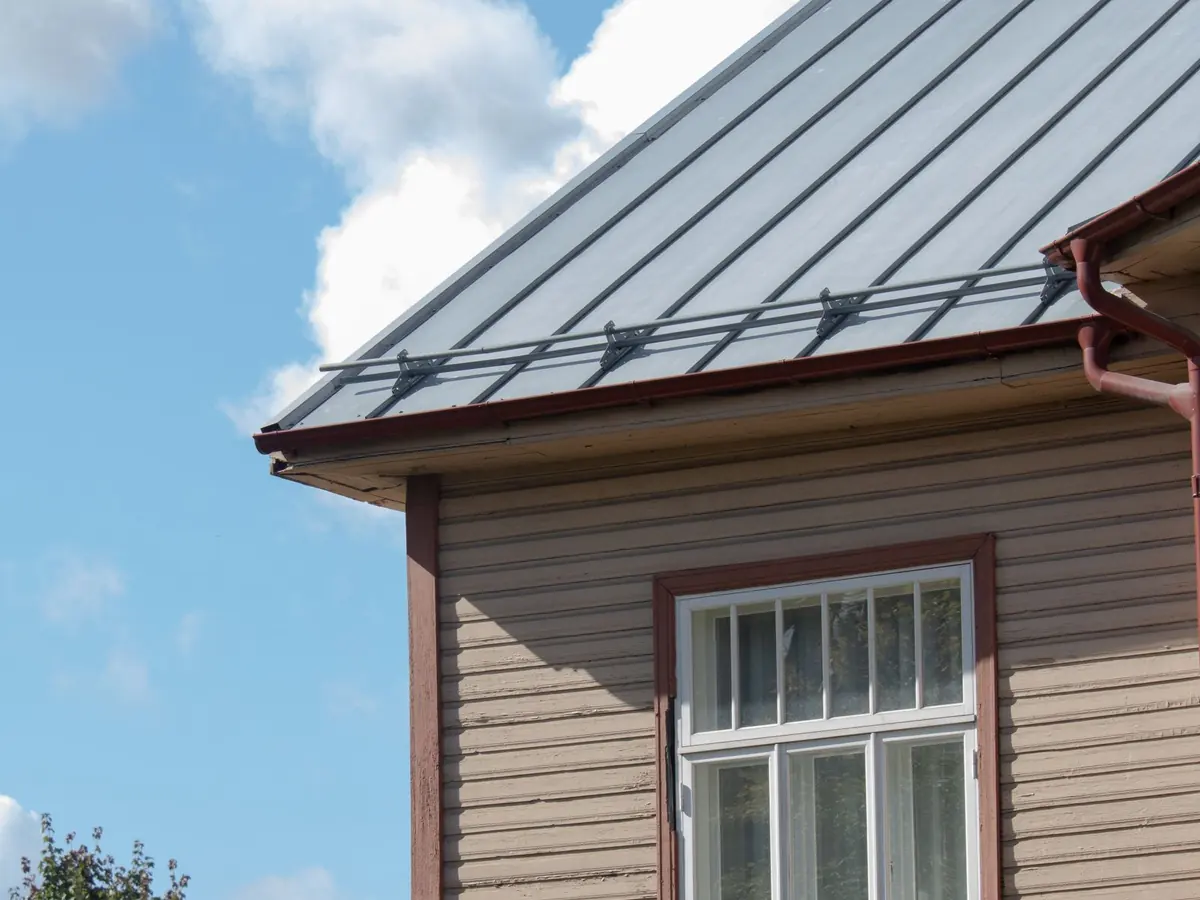 Gray standing-seam metal roof with raised vertical seams above a tan-sided house wall and a white-framed window