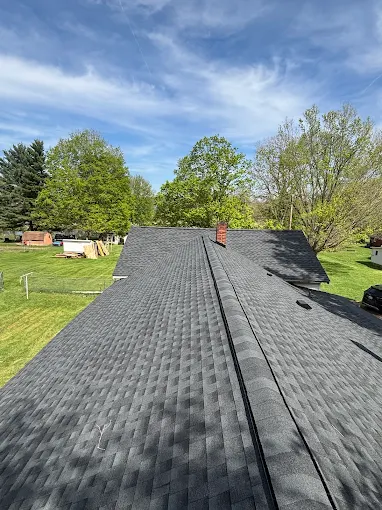Completed charcoal asphalt shingle roof with a continuous ridge vent, blue sky and spring trees behind it